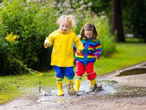 Two children wearing rain boots and jumping in puddles on the side of a street.
